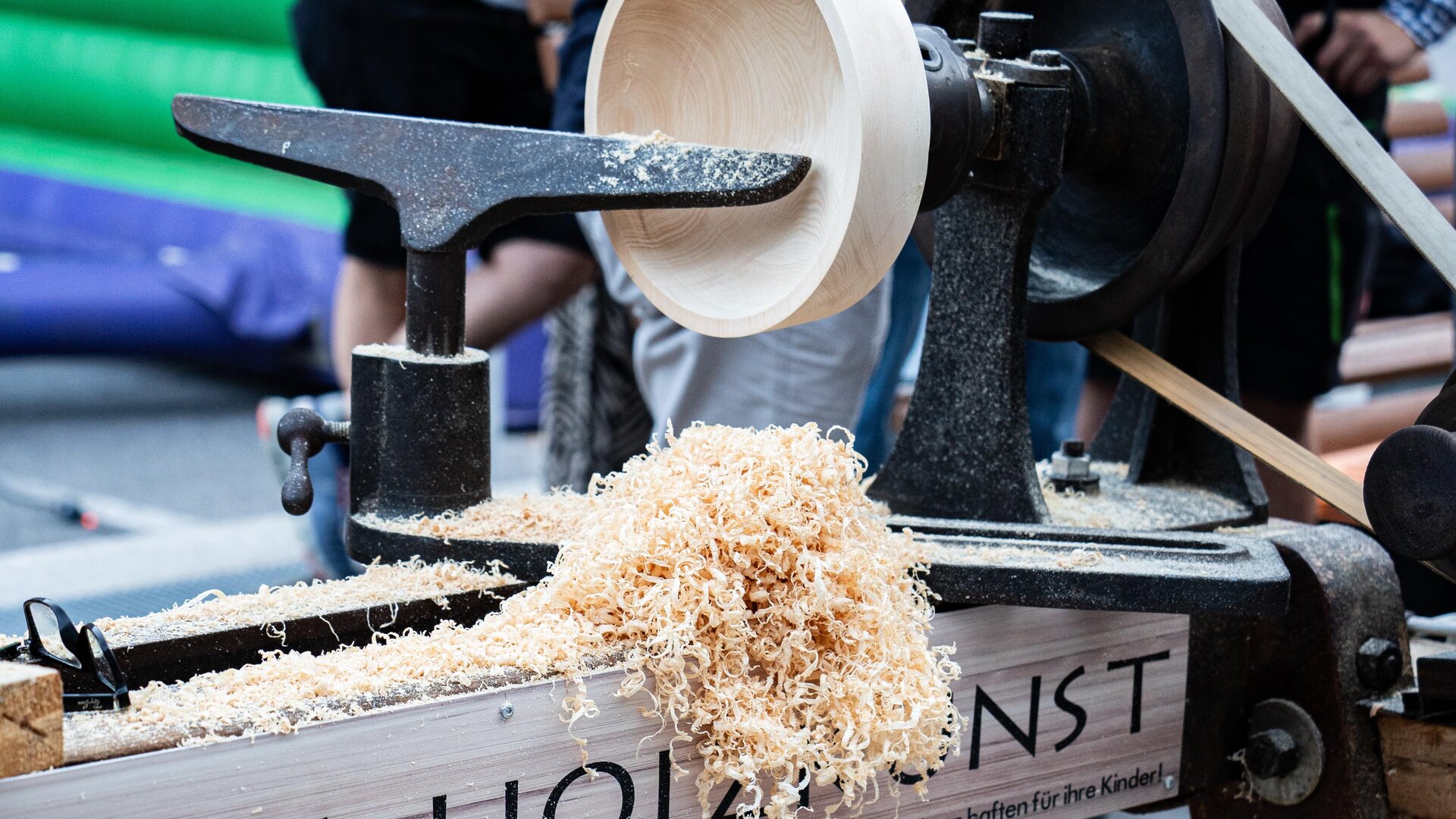 A wood lathe is used to turn a bowl. Shavings fall from the machine onto the floor.