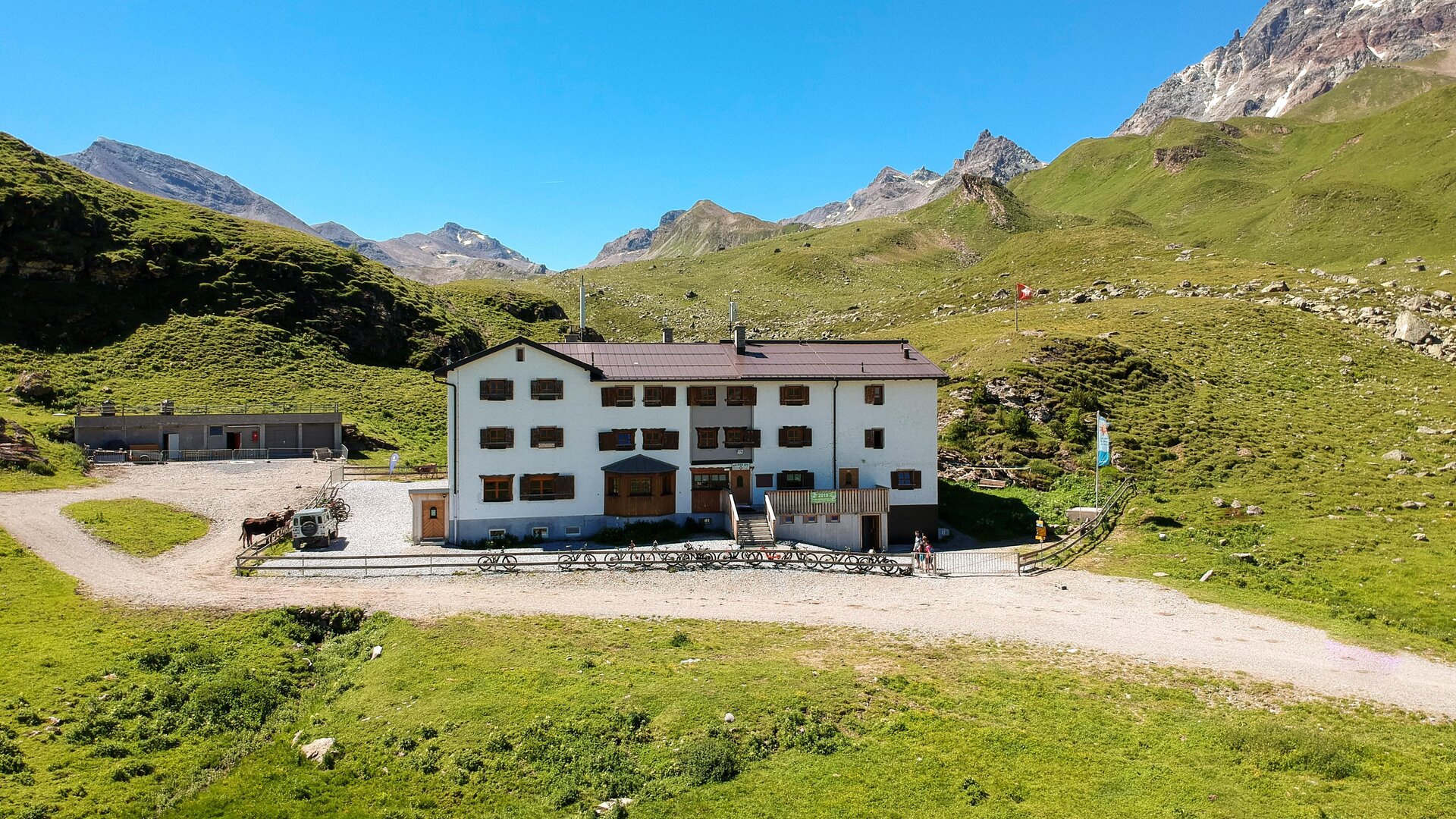 A mountain hut surrounded by green meadows and mountains. The sky is clear and blue.