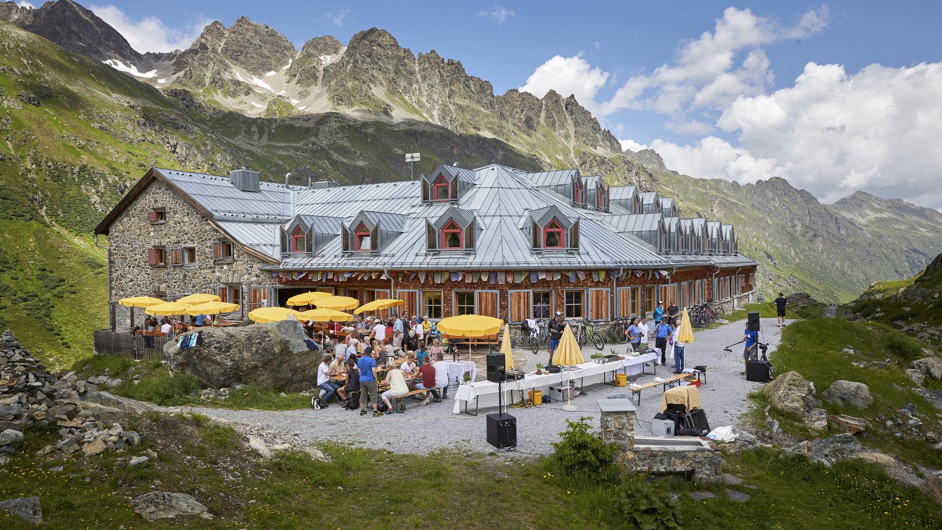 A charming mountain cabin with terraces and sun umbrellas. In the background, majestic mountains and a green meadow can be seen.