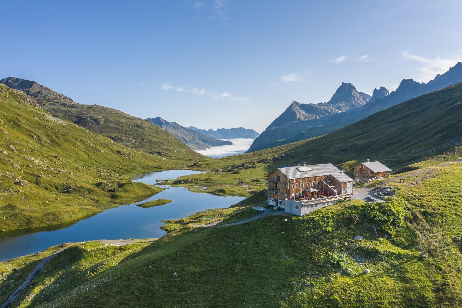 A modern wooden building is situated picturesque in the mountains. In the background, snow-covered peaks and a green landscape can be seen.