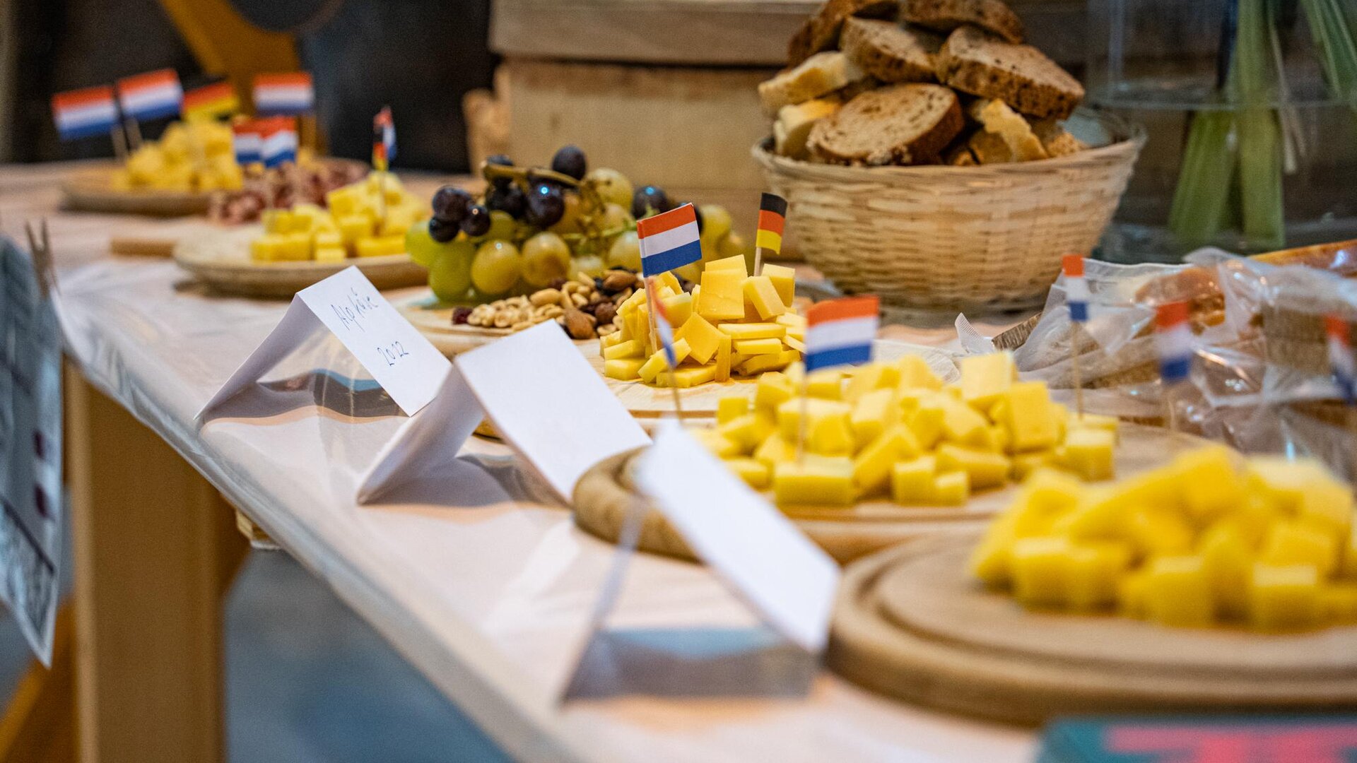 Eine schön dekorierte Käseplatte mit verschiedenen Käsesorten und frischem Brot. Auf dem Tisch sind kleine Flaggen und Obst zu sehen.