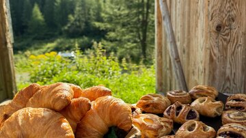 Ein Korb voller frisch gebackener Croissants steht neben einer Platte mit verschiedenen Gebäckstücken. Im Hintergrund sind grüne Bäume und Berge zu sehen.