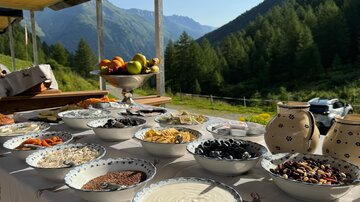 Ein Tisch gedeckt mit verschiedenen Schalen voller Zutaten in der Natur. Die Berge und der klare Himmel im Hintergrund schaffen eine gemütliche Atmosphäre.