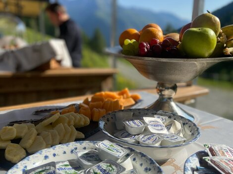 Ein schön gedeckter Tisch mit verschiedenen Lebensmitteln, darunter frisches Obst und Frischkäseportionen. Im Hintergrund sind klare Berge und ein blauer Himmel zu sehen.