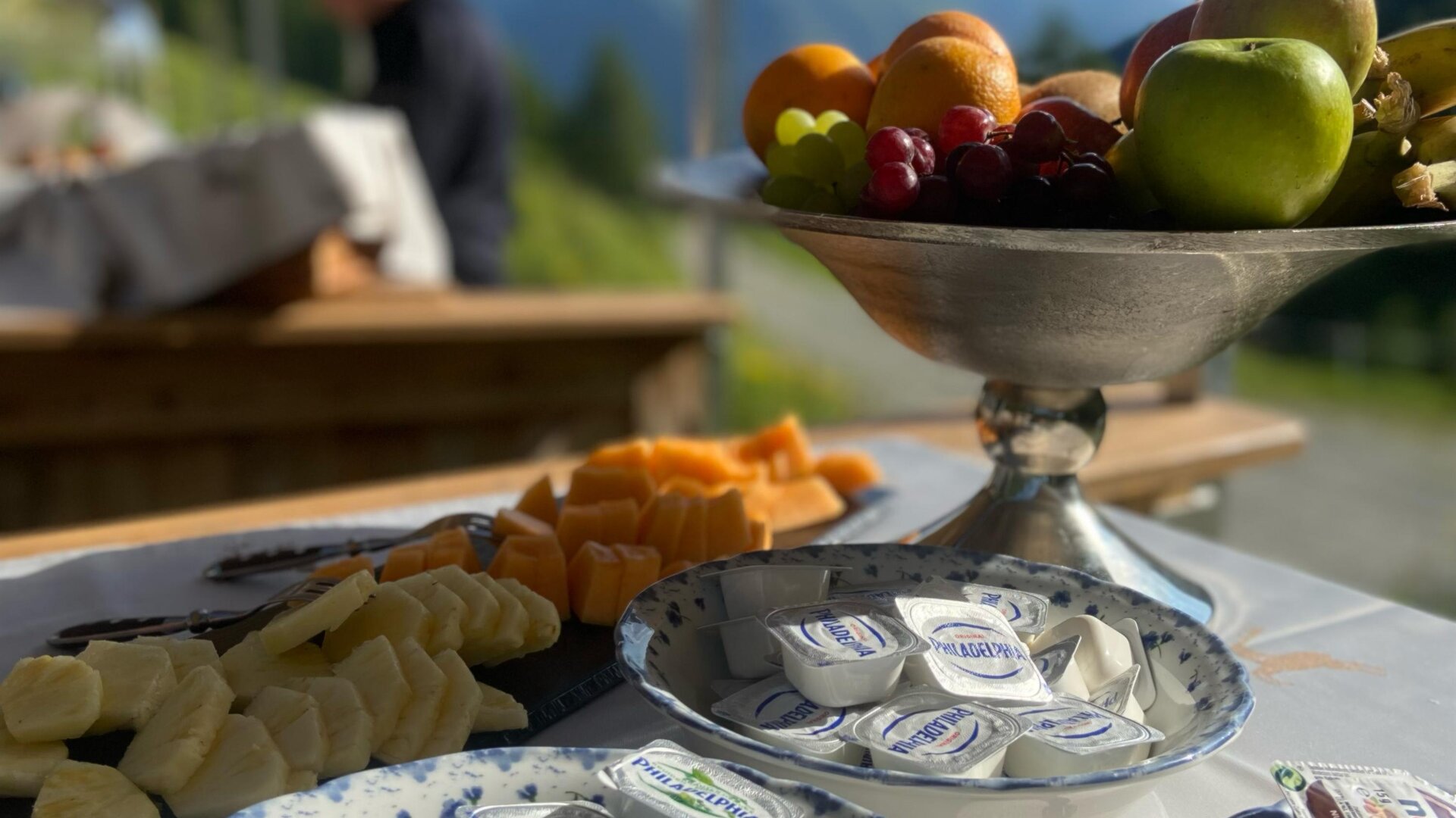 Ein schön gedeckter Tisch mit verschiedenen Lebensmitteln, darunter frisches Obst und Frischkäseportionen. Im Hintergrund sind klare Berge und ein blauer Himmel zu sehen.