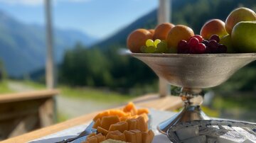 Ein rustikaler Tisch mit verschiedenen Käsesorten und frischem Obst. Im Hintergrund sind Berge und ein klarer blauer Himmel zu sehen.