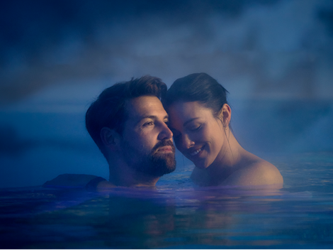 A couple stands in calm water, surrounded by steam. They smile at each other and appear relaxed.