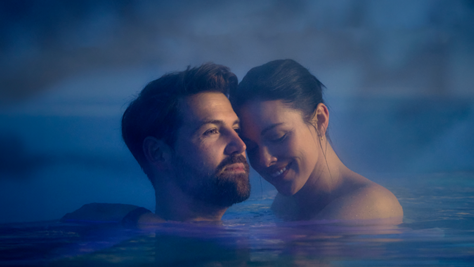 A couple stands in calm water, surrounded by steam. They smile at each other and appear relaxed.