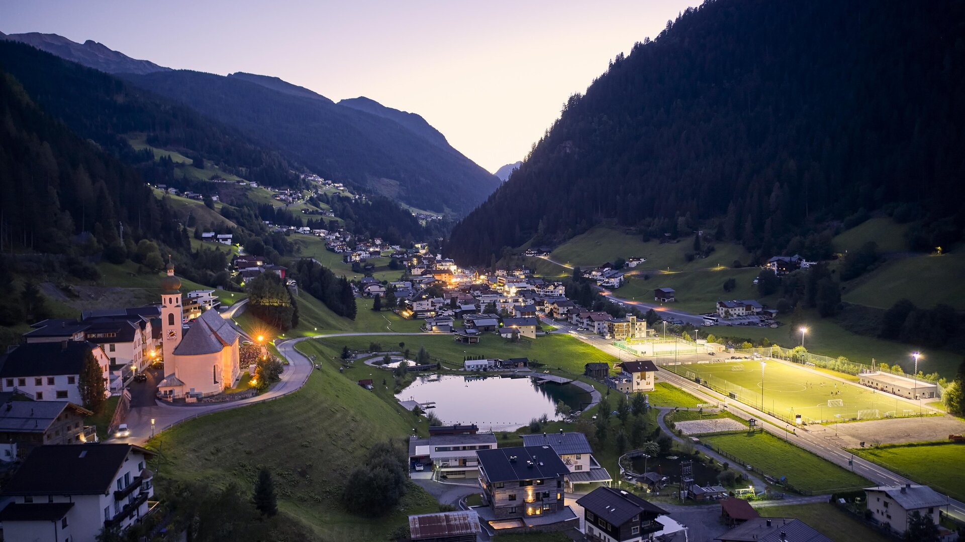 A picturesque mountain landscape with a serene valley and a small village. The surroundings are lit at dusk, showcasing a peaceful atmosphere.