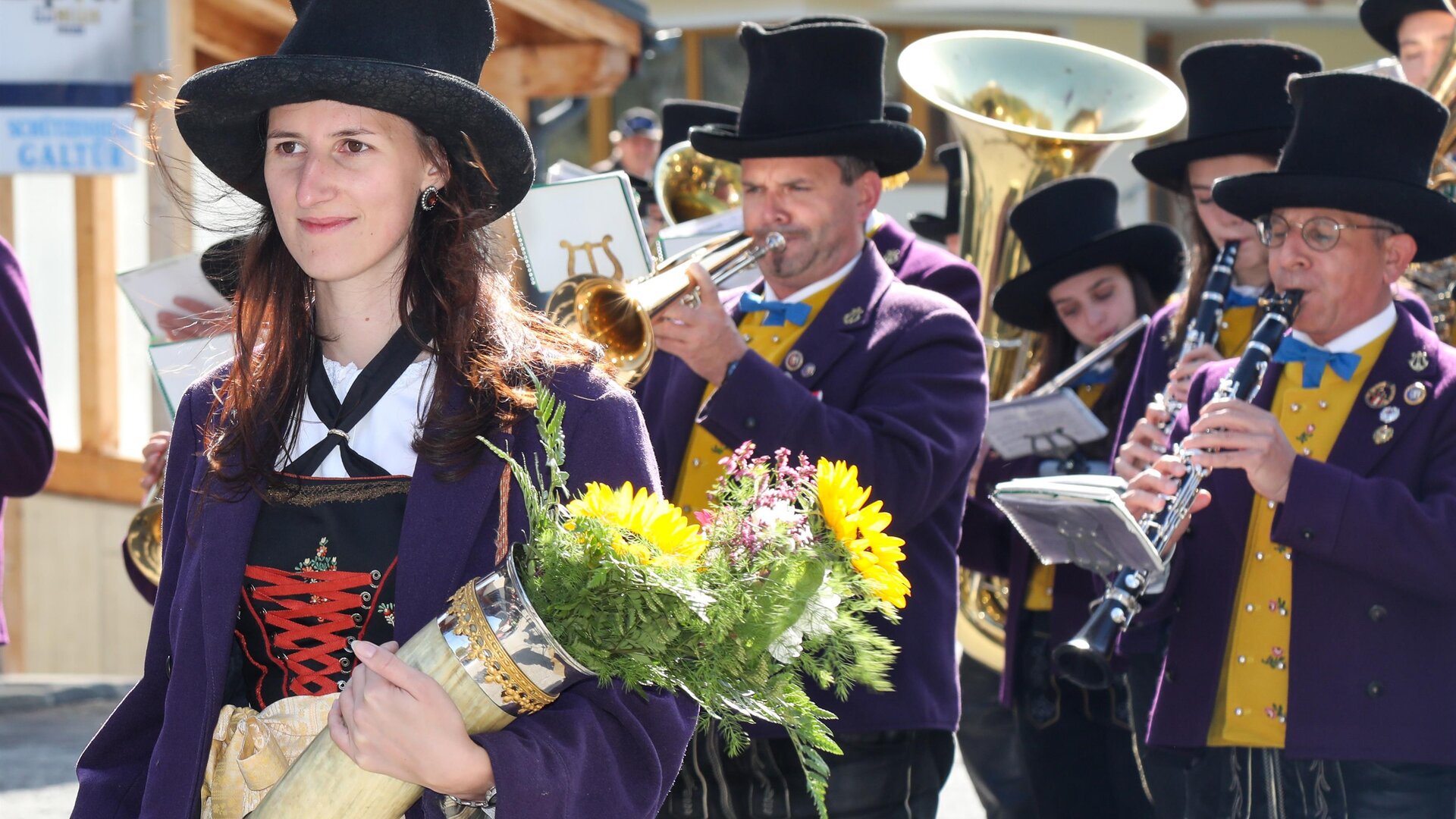 Eine Gruppe von Menschen in traditionellen Trachten marchiert durch die Stadt. Eine Frau hält einen Strauß Blumen in der Hand.