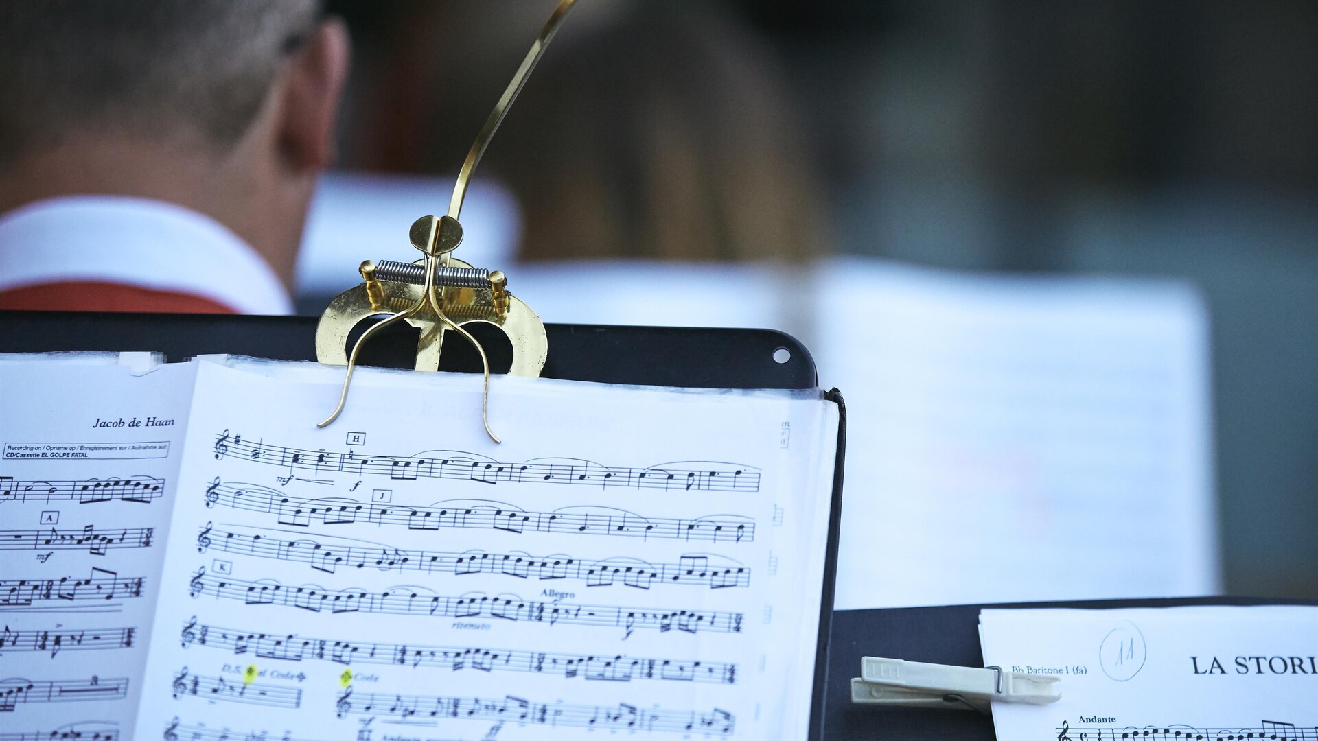 Ein Ausschnitt von Notenblättern auf einem Notenständer. Im Hintergrund sind Musiker zu sehen, die sich auf eine Aufführung vorbereiten.