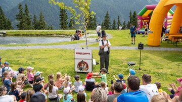 Eine große Gruppe von Kindern sitzt auf dem Gras und schaut einem Performer zu. Im Hintergrund sind eine aufblasbare Hüpfburg und schöne Berge zu sehen.