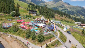 Ein lebhaftes Bergtal mit vielen Bäumen und einer bunten Terrasse voller Sonnenschirme. Im Hintergrund sind majestätische Berge und ein strahlend blauer Himmel zu sehen.