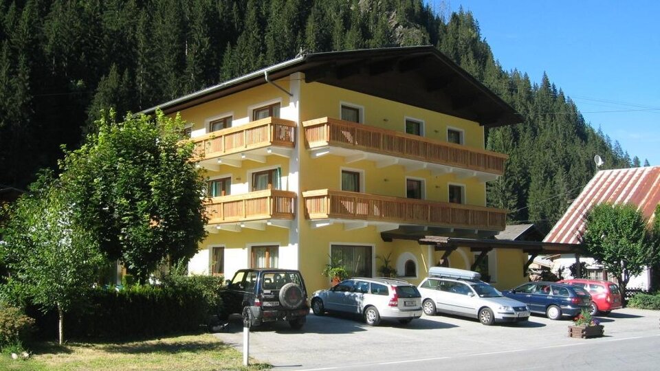 A bright building with several balconies, surrounded by trees and mountains. Several cars are parked in the parking lot.