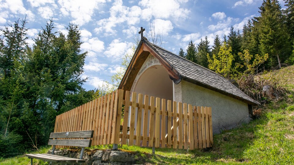 A small chapel with a wooden fence stands in a wooded environment. The sky is partly cloudy and the landscape appears calm and peaceful. | © Bernhard Gruber – Photography