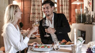 Ein romantisches Abendessen in einem warmen, holzverkleideten Raum. Ein Paar stößt mit Weingläsern an, umgeben von einem eleganten Tisch mit köstlichem Essen.