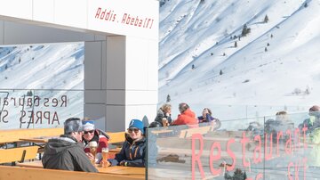 A group of people is seated in an outdoor restaurant, surrounded by snow-covered mountains. The sun is shining, and there is a relaxed atmosphere.