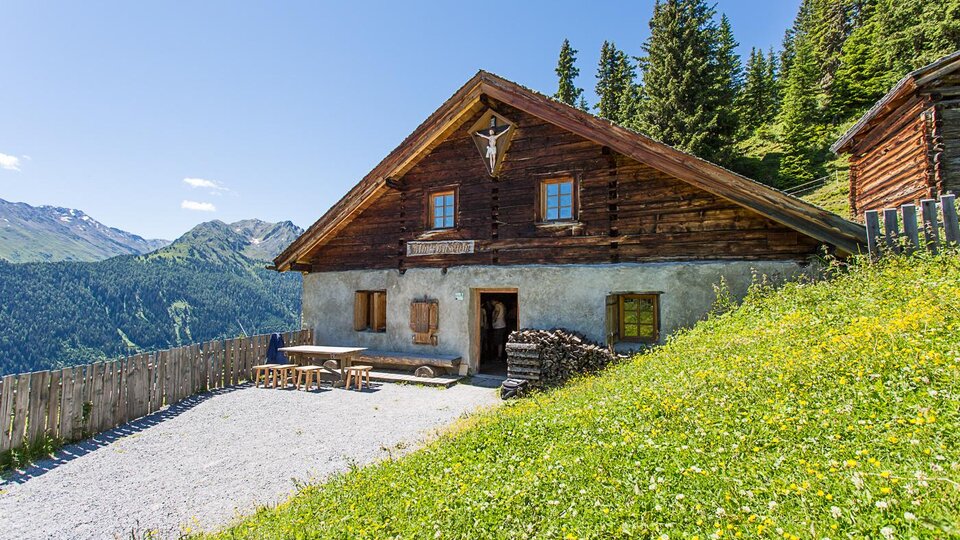 A traditional wooden house in the mountains, surrounded by green meadows and trees. The clear, sunny landscape offers a beautiful view of the surrounding mountains.