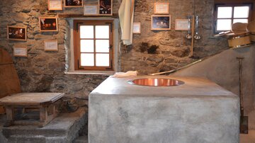 A rustic kitchen with a large, round copper container and stone walls. Pictures and documents hang on the walls.