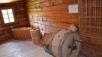 A rustic room with wooden walls, equipped with a large wooden barrel and two other containers. The light falls through a window onto the wooden objects.