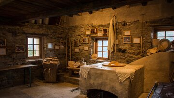 A cozy and rustic interior view of an old farmhouse. Visible are wooden and stone structures as well as agricultural tools.