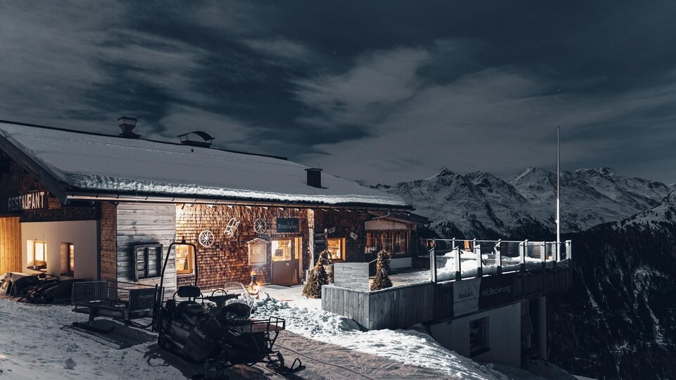 A wooden ski hut in the snow with a breathtaking mountain panorama. The scene is at night with a clear sky and a lit interior.
