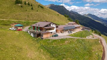A cozy mountain cabin surrounded by green meadows and mountains. The sky is clear with a few clouds.