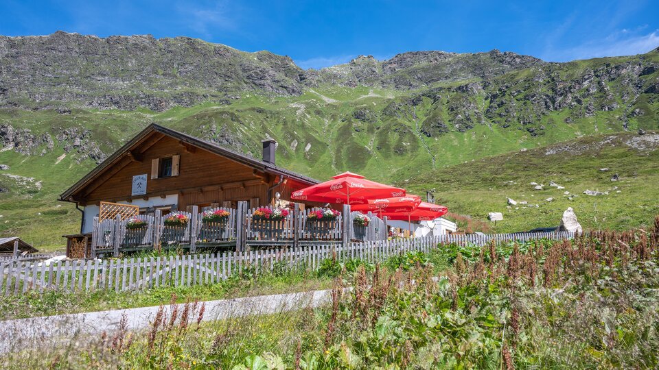A picturesque mountain hut surrounded by green meadows and mountains. Red sunshades stand in front of the terrace, which looks inviting.