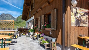 A charming mountain cabin with wooden walls and blooming flowers. Outside, tables and benches are set up on a wooden terrace.