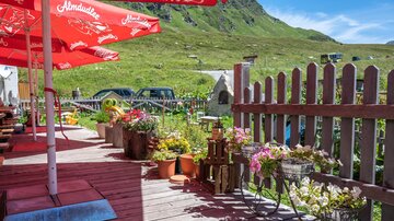 A cozy outdoor area with red umbrellas and blooming plants. In the background, green mountains and some cars can be seen.