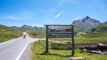 A picturesque mountain landscape with a clear blue sky. A wooden sign points to a hiking trail.