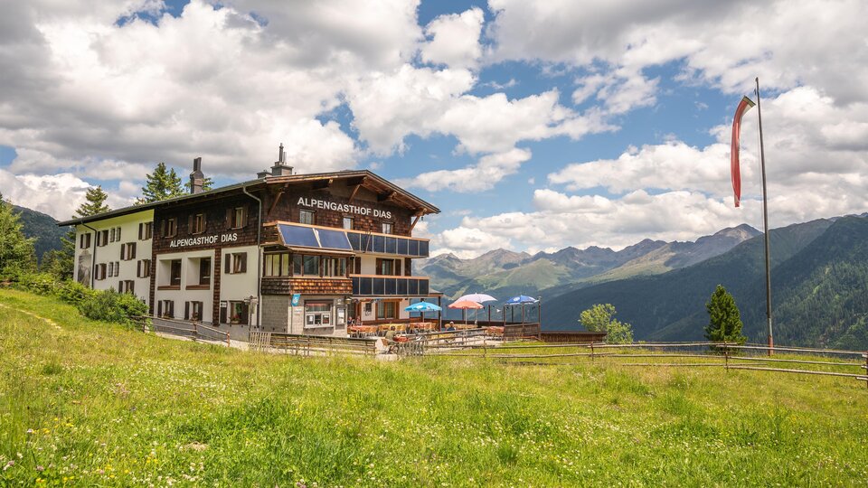 A picturesque mountain cabin surrounded by green meadows and mountains. The sky is partly cloudy and the atmosphere is inviting.