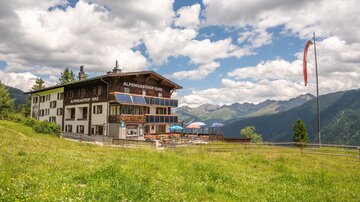 A picturesque mountain cabin surrounded by green meadows and mountains. The sky is partly cloudy and the atmosphere is inviting.