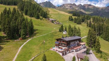 A picturesque mountain panorama with plenty of greenery and clouds in the sky. In the foreground, there is a cozy hotel surrounded by forests and hiking trails.