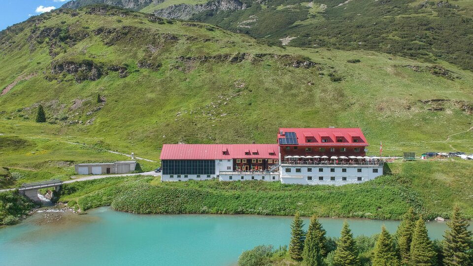 Ein malerisches Hotel am Ufer eines klaren, blauen Sees, umgeben von grünen Hügeln. Im Hintergrund sind Berge und ein blauer Himmel zu sehen.