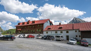 Ein großes Gebäude mit rotem Dach in einem bergigen Gebiet. Auf dem Parkplatz stehen mehrere Autos und der Himmel ist blau mit weißer Bewölkung. | © TVB Paznaun - Ischgl