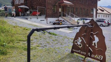 Ein rustikales Gebäude mit einer Holzfassade, das als Unterkunft dient. Im Vordergrund befindet sich ein Hinweisschild für Radfahrer und Wanderer.
