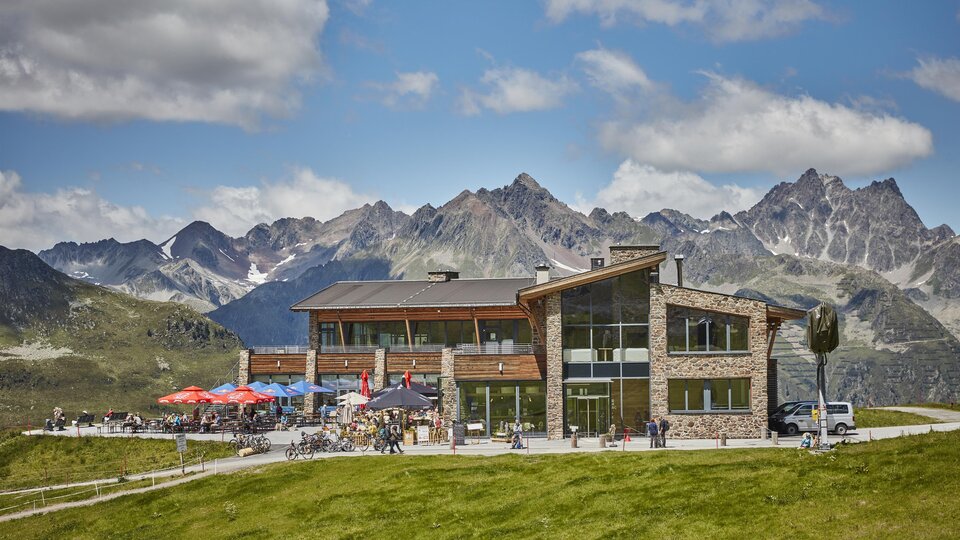 A modern building in a picturesque mountain landscape. The terrace is adorned with guests and colorful umbrellas.
