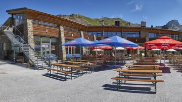 A modern alpine guesthouse with a terrace and several tables. In the background, mountains and a clear sky can be seen.