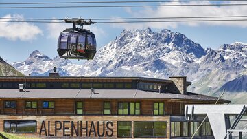 A cable car travels over the Alpine house in front of impressive mountain peaks. The landscape showcases the beauty of the Alps with clear skies and snow-covered mountains.