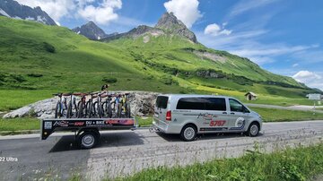 A vehicle with a trailer full of bicycles drives through a green mountain landscape. The surroundings are sunny with impressive mountains in the background.