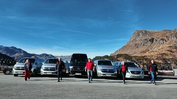A group of people is standing in front of several cars in a mountainous landscape. The sky is blue and the surroundings are sunny.