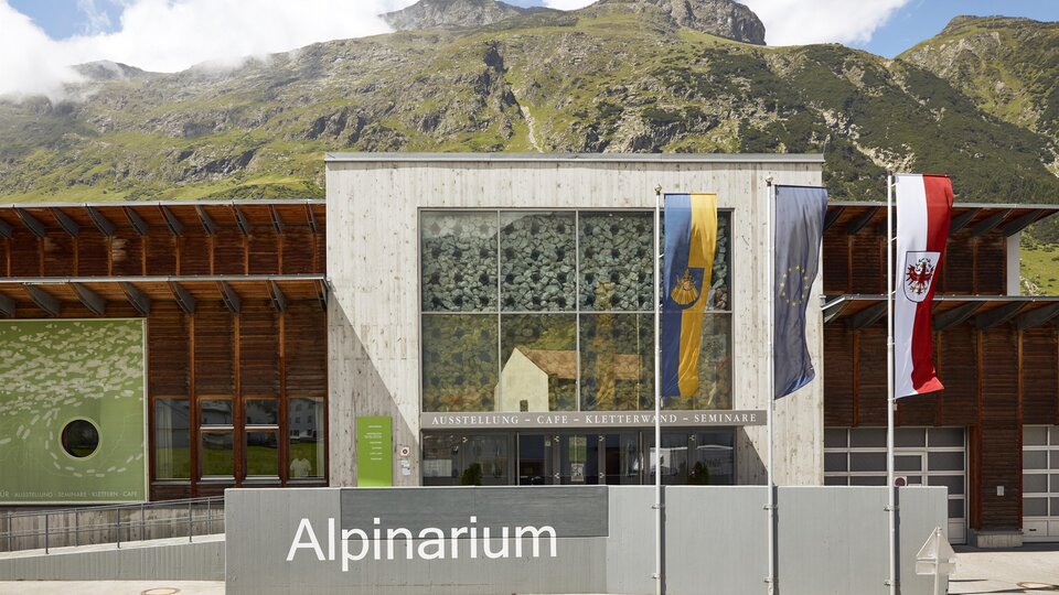 A modern building of the Alpinarium with large windows. In the background, green mountains and a blue sky can be seen.