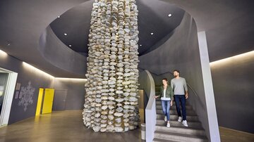 A modern interior with a striking stone column and a curved staircase. Two people are walking down the staircase. | © TVB Paznaun - Ischgl