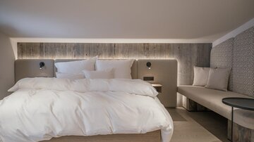 A modern bedroom with a large bed and white bedding. A cozy seating area and soft lighting create a relaxing atmosphere.