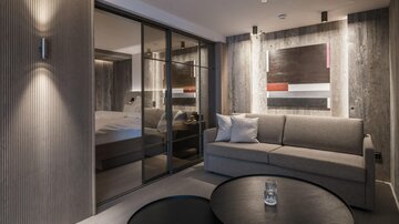 A modern room with gray walls and a comfortable sofa. In the background, an elegant bedroom is visible.