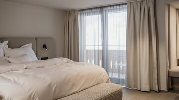 A modern bedroom with a large bed and plenty of natural light.  
Large windows with curtains provide a view outside.