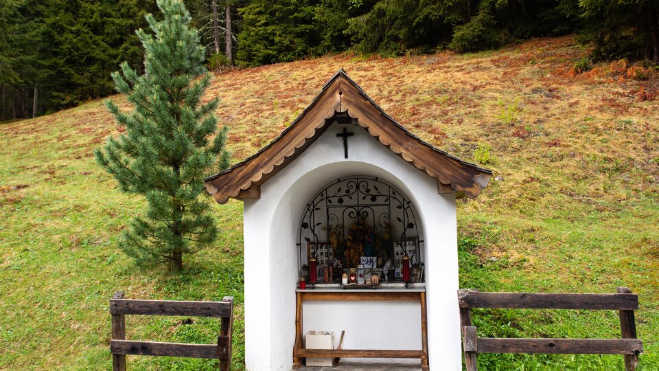 A small wooden shrine stands in a green field, surrounded by trees. The shrine has a red roof and is decorated with a cross and small statues. | © Bernhard Gruber – Photography
