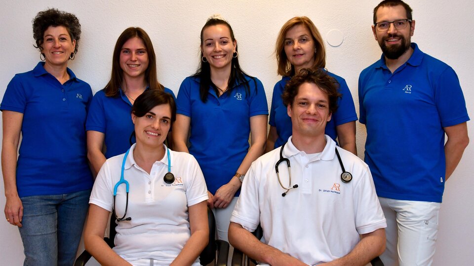 Ein Team von Fachleuten steht zusammen in blauen T-Shirts. Zwei Personen tragen Stethoskope und lächeln freundlich in die Kamera.