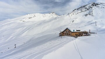 Eine Berghütte im Schnee, umgeben von schneebedeckten Bergen. Die Landschaft wirkt ruhig und winterlich.